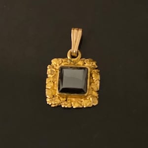 Op de afbeelding: Een vierkante hanger met een centrale zwarte edelsteen in een getextureerd goudkleurig frame met een grillige, organische rand. De hanger is voorzien van een klein goudkleurig oogje aan de bovenkant voor bevestiging aan een ketting, wat een contrast laat zien tussen de gladde donkere steen en de ruwe, metalen rand.