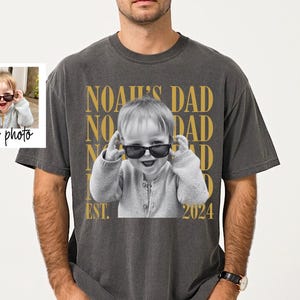 May include: Dark gray t-shirt featuring a black and white photo of a child wearing sunglasses. The text "NOAH'S DAD" is printed in gold, along with "EST. 2024". A small photo is in the upper left corner with the text "your photo".