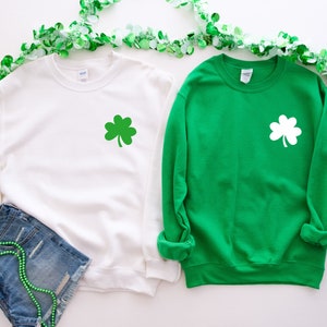 Puede incluir: Dos sudaderas con un dise&ntilde;o de tr&eacute;bol. Una sudadera es blanca con un tr&eacute;bol verde y la otra es verde con un tr&eacute;bol blanco.