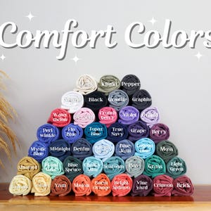 Op de afbeelding: Een piramide van opgerolde t-shirts in verschillende kleuren, waaronder wit, zwart en blauw. De tekst "Comfort Colors" staat boven de shirts. De shirts zijn gerangschikt op een houten oppervlak.