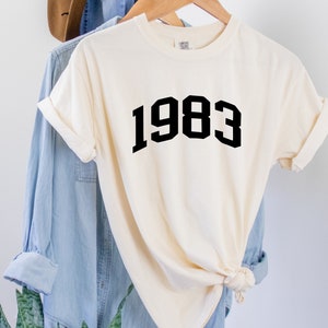May include: A white t-shirt with black text that reads "1983" in a bold, blocky font.