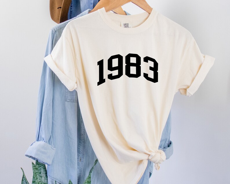 May include: A white t-shirt with black text that reads "1983" in a bold, blocky font.