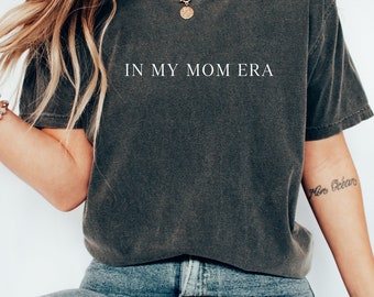 May include: A dark gray t-shirt with the text "IN MY MOM ERA" printed in white on the front. The shirt is being worn by a person with long blonde hair and a gold necklace.