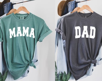 Peut inclure: Deux t-shirts, un vert sarcelle avec "MAMA" en texte blanc et un gris avec "DAD" en texte blanc. Les deux t-shirts sont suspendus à des cintres.