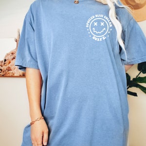 Puede incluir: Una camiseta azul claro con un gr&aacute;fico blanco de una cara sonriente con X para los ojos. El gr&aacute;fico est&aacute; dentro de un c&iacute;rculo con el texto "Spoiled Blue Collar Wife" alrededor.