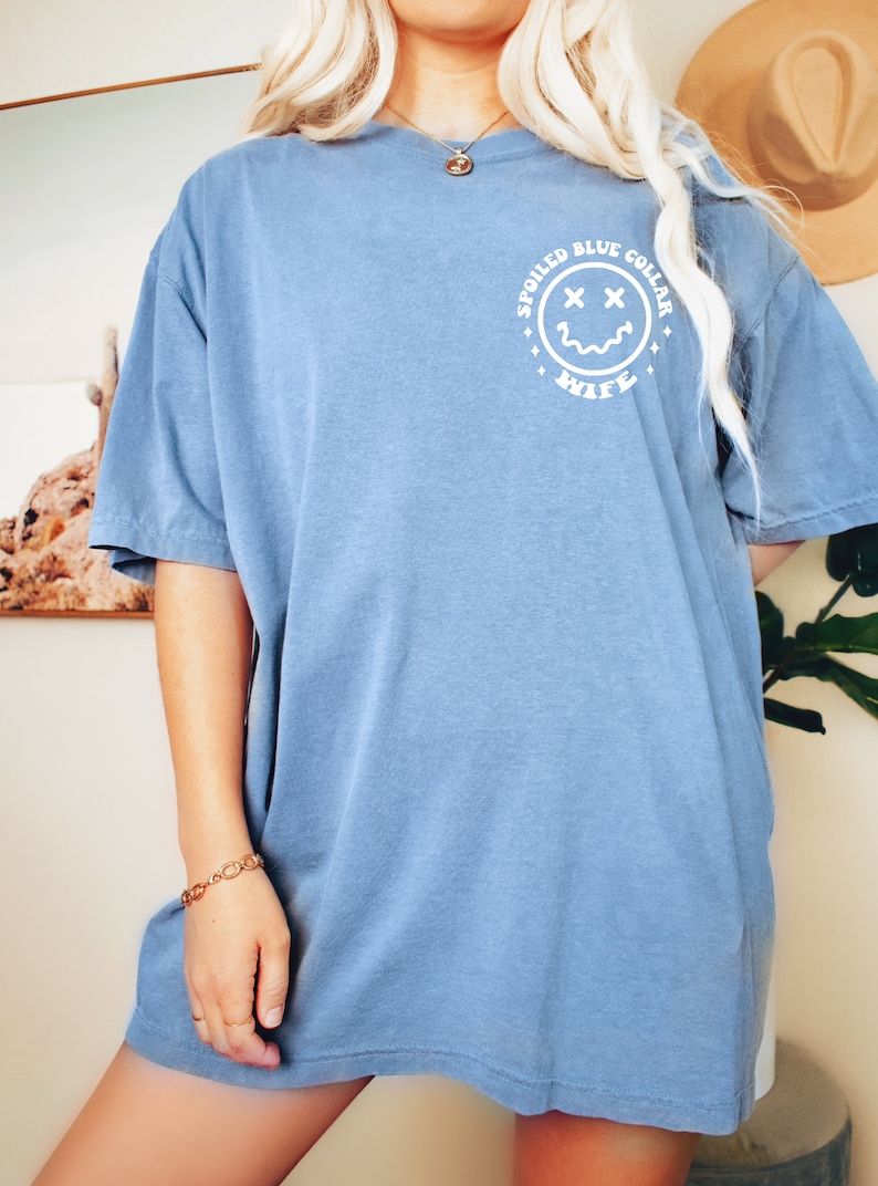 Puede incluir: Una camiseta azul claro con un gr&aacute;fico blanco de una cara sonriente con X para los ojos. El gr&aacute;fico est&aacute; dentro de un c&iacute;rculo con el texto "Spoiled Blue Collar Wife" alrededor.