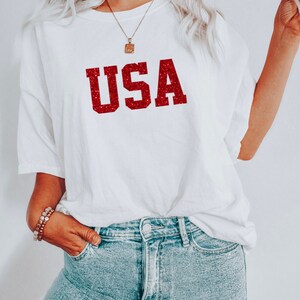 May include: White t-shirt with a red glitter "USA" graphic. The shirt is oversized and has a relaxed fit.