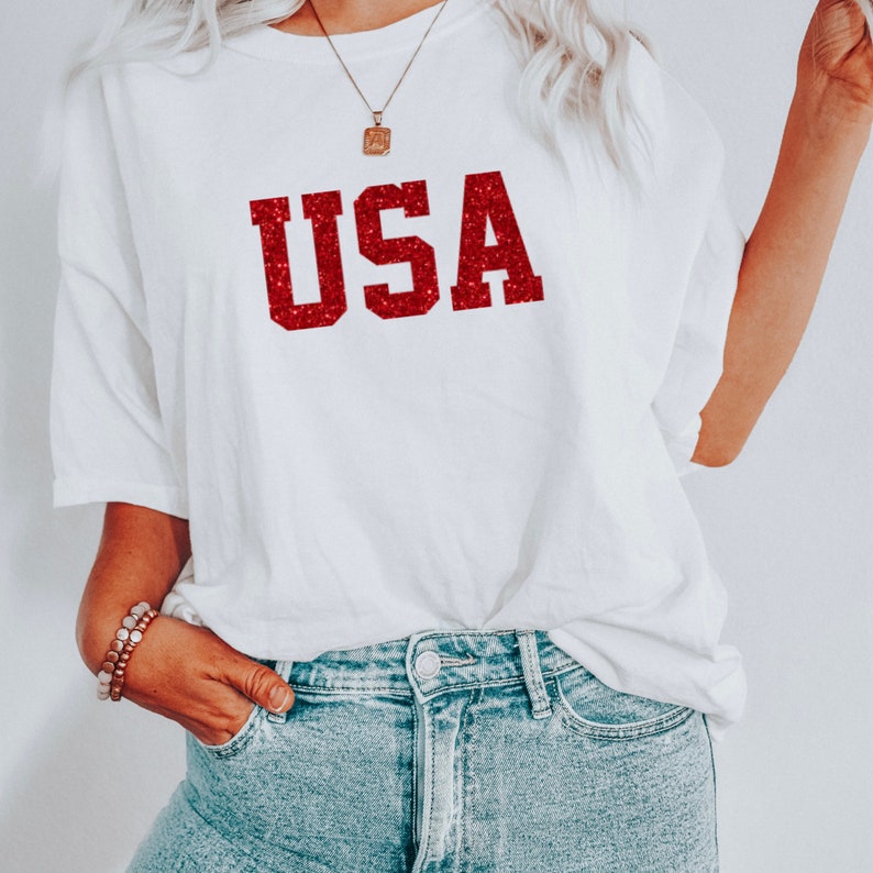 May include: White t-shirt with a red glitter "USA" graphic. The shirt is oversized and has a relaxed fit.