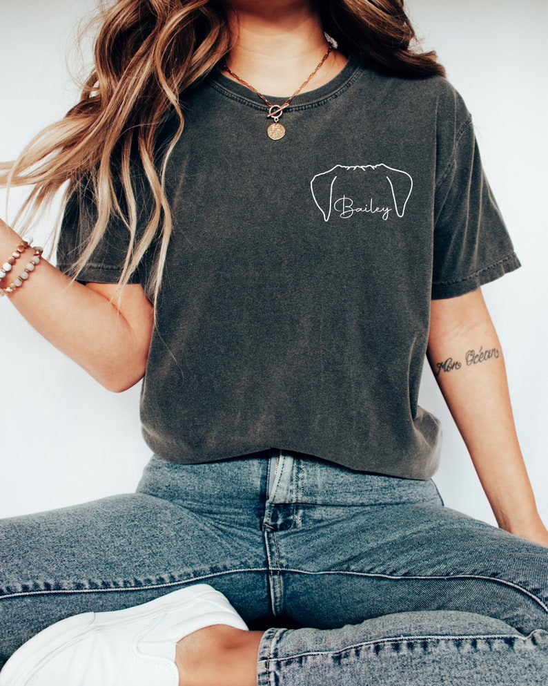 Puede incluir: Una camiseta gris oscuro con un contorno blanco de la cabeza de un perro y el nombre "Bailey" escrito debajo. La camiseta la lleva puesta una persona con el pelo largo y casta&ntilde;o y un collar de oro.