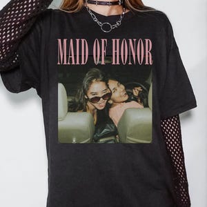 Op de afbeelding: Zwart T-shirt met de tekst "MAID OF HONOR" in roze. Het shirt heeft een foto van twee vrouwen in een auto. De mouwen zijn lang en de persoon draagt een zwarte choker-ketting.