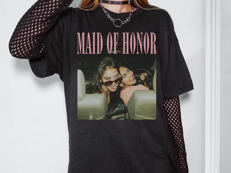 Op de afbeelding: Zwart T-shirt met de tekst "MAID OF HONOR" in roze. Het shirt heeft een foto van twee vrouwen in een auto. De mouwen zijn lang en de persoon draagt een zwarte choker-ketting.