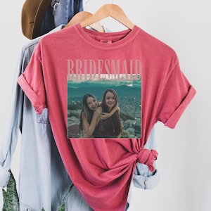 Op de afbeelding: Een rood t-shirt met het woord "BRIDESMAID" in een lichtroze lettertype. Het shirt heeft een foto van twee vrouwen die elkaar omarmen. Het shirt hangt aan een houten hanger met andere kledingstukken op de achtergrond.
