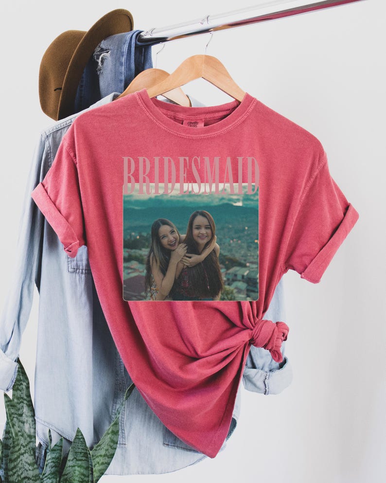 Op de afbeelding: Een rood t-shirt met het woord "BRIDESMAID" in een lichtroze lettertype. Het shirt heeft een foto van twee vrouwen die elkaar omarmen. Het shirt hangt aan een houten hanger met andere kledingstukken op de achtergrond.