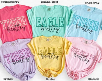 May include: A collection of six short-sleeved t-shirts in various colors, including pink, light blue, yellow, and purple. Each shirt features a team name and the name "Bentley" in a collegiate font.