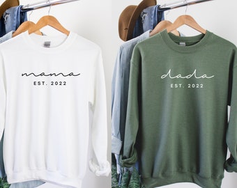 Puede incluir: Dos sudaderas, una blanca y otra verde, con las palabras "mama est. 2022" y "dada est. 2022" impresas en ellas.