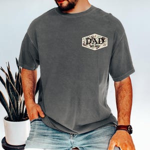 Puede incluir: Una camiseta gris carb&oacute;n con un parche de camuflaje que dice "DAD EST. 2023". La camiseta es de corte holgado y se combina con vaqueros azules. Se ve un reloj de cuero marr&oacute;n en la mu&ntilde;eca.