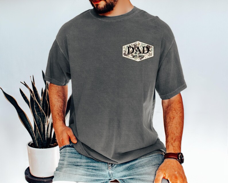 Puede incluir: Una camiseta gris carb&oacute;n con un parche de camuflaje que dice "DAD EST. 2023". La camiseta es de corte holgado y se combina con vaqueros azules. Se ve un reloj de cuero marr&oacute;n en la mu&ntilde;eca.