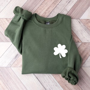 Puede incluir: Una sudadera verde con un dise&ntilde;o de tr&eacute;bol blanco. La sudadera est&aacute; doblada y colocada sobre una superficie de madera.