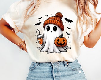 May include: A white t-shirt with a cartoon ghost wearing an orange and brown knit hat, holding a pumpkin and a cup of orange beverage. The ghost has large eyes and a surprised expression. There are bats and stars in the background.
