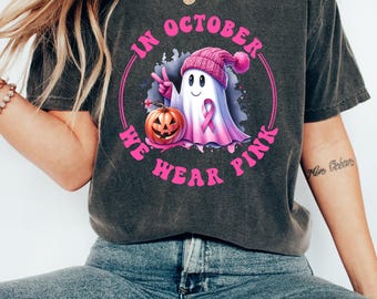 May include: A gray t-shirt with a pink and white ghost wearing a pink hat and holding a pumpkin. The text "In October We Wear Pink" is in a circle around the ghost.