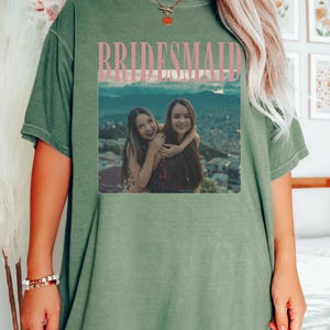 Op de afbeelding: Olijfgroen T-shirt met het woord "BRIDESMAID" in roze blokletters boven een foto van twee vrouwen. Het woord "MOSS" staat linksboven gedrukt. Het shirt heeft een relaxte pasvorm.