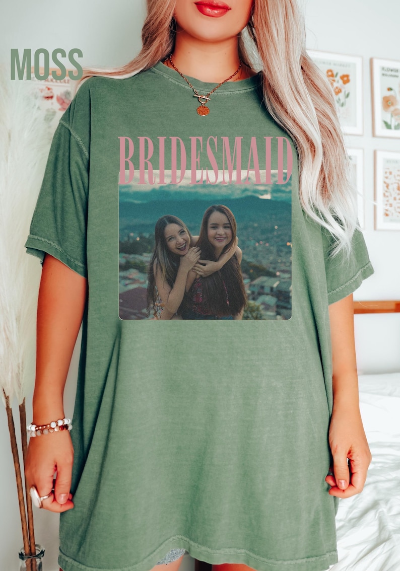 Op de afbeelding: Olijfgroen T-shirt met het woord "BRIDESMAID" in roze blokletters boven een foto van twee vrouwen. Het woord "MOSS" staat linksboven gedrukt. Het shirt heeft een relaxte pasvorm.
