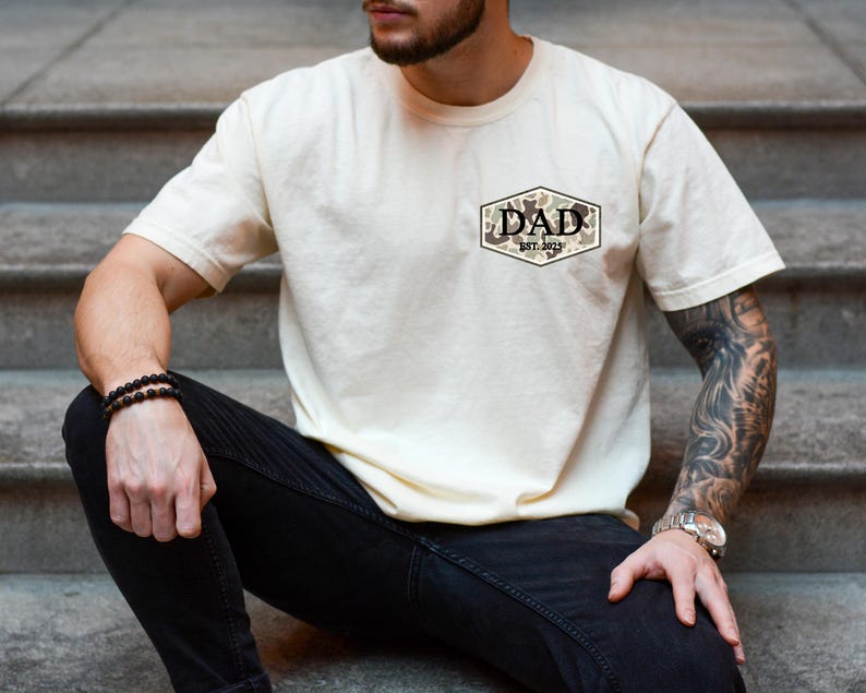 Puede incluir: Camiseta color crema con un dise&ntilde;o de camuflaje y la palabra "DAD" dentro de un hex&aacute;gono marr&oacute;n y beige. El texto "EST. 2023" est&aacute; debajo de la palabra "DAD". La camiseta es de manga corta.