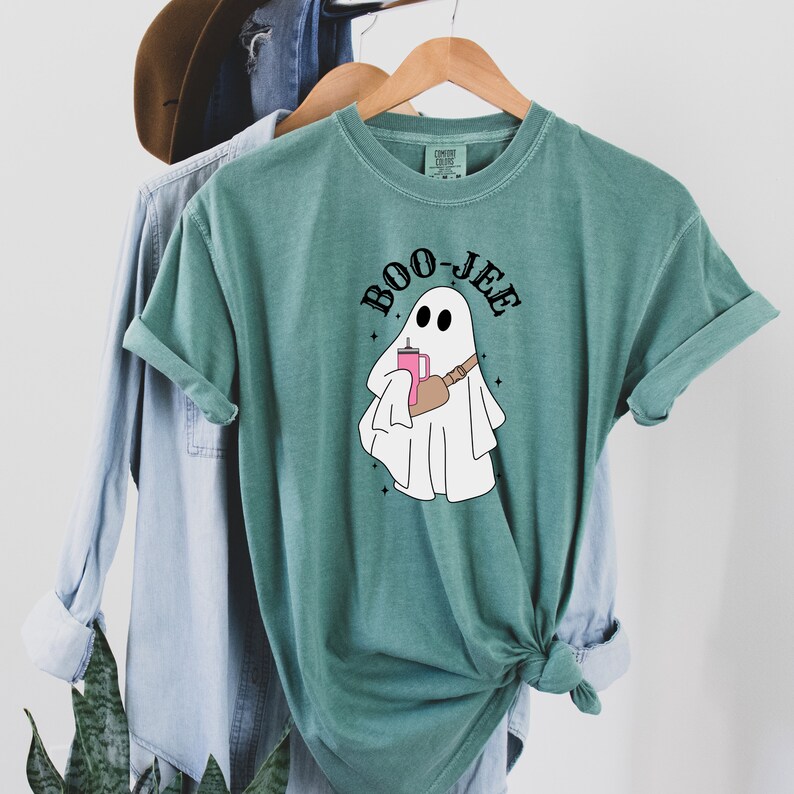 May include: A teal green t-shirt with a white ghost holding a pink drink and a black strap across its body. The text "BOO-JEE" is written in black above the ghost.