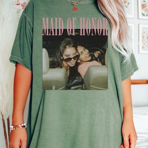 Op de afbeelding: Olijfgroen t-shirt met het woord "MOSS" in de linkerbovenhoek. Het shirt heeft een foto van twee vrouwen in een auto met de woorden "MAID OF HONOR" in roze. Het t-shirt heeft een relaxte pasvorm.
