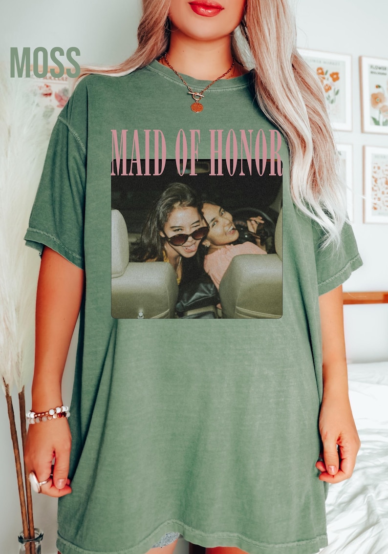 Op de afbeelding: Olijfgroen t-shirt met het woord "MOSS" in de linkerbovenhoek. Het shirt heeft een foto van twee vrouwen in een auto met de woorden "MAID OF HONOR" in roze. Het t-shirt heeft een relaxte pasvorm.
