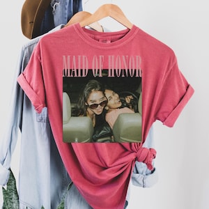 Op de afbeelding: Een rood t-shirt met de tekst "MAID OF HONOR" bovenaan. Daaronder een foto van twee vrouwen in een auto. Het shirt hangt aan een houten hanger met andere kledingstukken op de achtergrond.