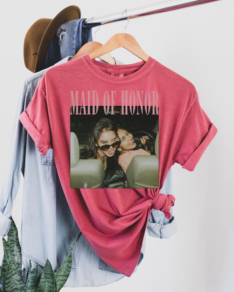 Op de afbeelding: Een rood t-shirt met de tekst "MAID OF HONOR" bovenaan. Daaronder een foto van twee vrouwen in een auto. Het shirt hangt aan een houten hanger met andere kledingstukken op de achtergrond.