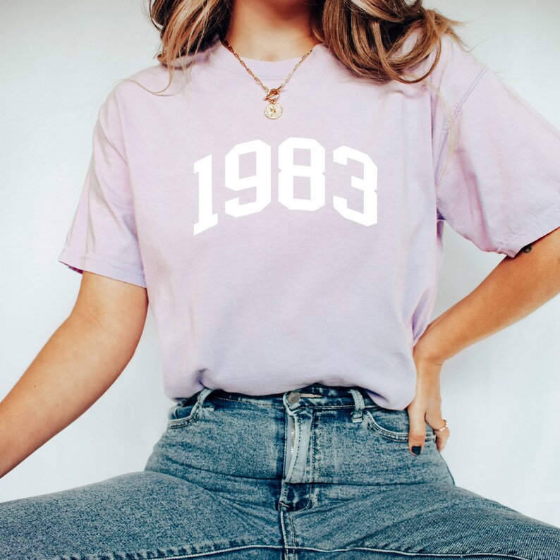 May include: A light purple t-shirt with the year 1983 printed in white on the front.