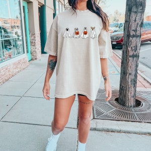 May include: A beige oversized t-shirt with a graphic of four dogs dressed as ghosts for Halloween. The dogs are wearing white sheets with faces peeking out. One dog is holding a pumpkin bucket.