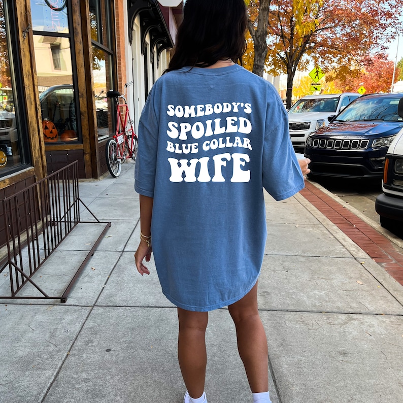 Puede incluir: Una camiseta azul de gran tama&ntilde;o con el texto "Somebody's Spoiled Blue Collar Wife" en blanco.