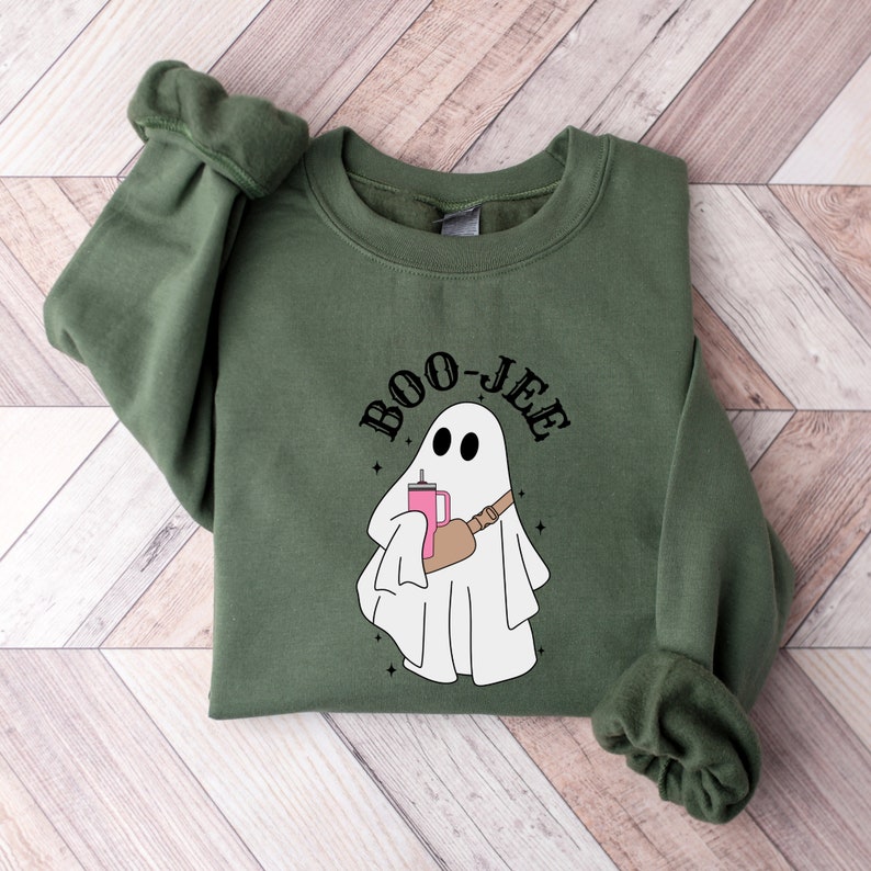 Pu&ograve; includere: Una felpa verde con un fantasma bianco che tiene una bevanda rosa e una borsa marrone. Il testo "BOO-JEE" &egrave; sopra il fantasma.