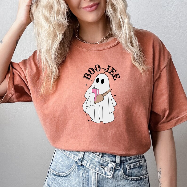 May include: A burnt orange t-shirt with a white ghost holding a pink drink and the text "BOO-JEE" in black.