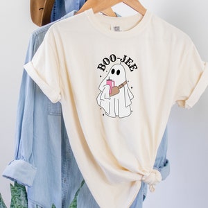 May include: A cream colored t-shirt with a black and white illustration of a ghost holding a pink drink and a brown bag. The text "BOO-JEE" is written in black above the ghost.