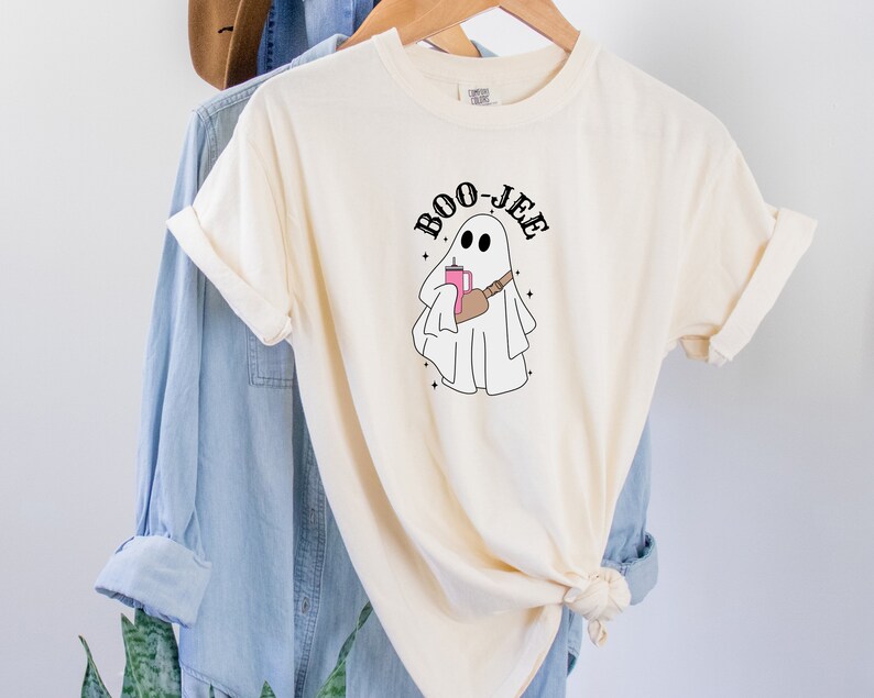 May include: A cream colored t-shirt with a black and white illustration of a ghost holding a pink drink and a brown bag. The text "BOO-JEE" is written in black above the ghost.