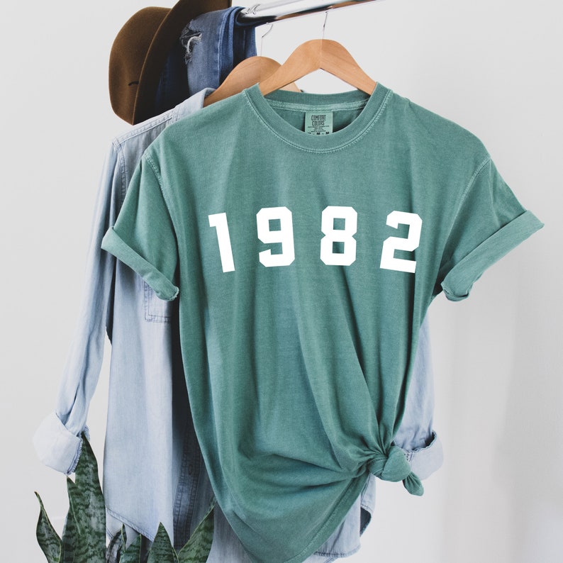 K&ouml;nnte beinhalten: Ein blaugr&uuml;nes T-Shirt mit dem Jahr 1982 in Wei&szlig; auf der Vorderseite.