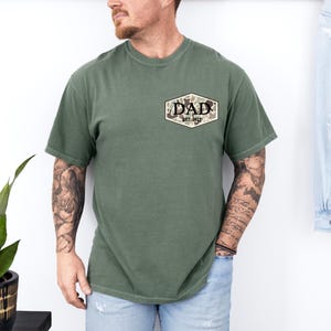 Puede incluir: Camiseta verde oliva con un bolsillo con un dise&ntilde;o de camuflaje y la palabra "DAD" con "EST. 2023" impresa debajo. La camiseta tiene mangas cortas y cuello redondo.