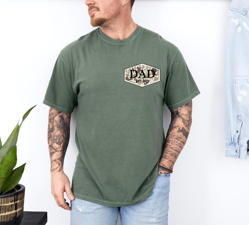 Puede incluir: Camiseta verde oliva con un bolsillo con un dise&ntilde;o de camuflaje y la palabra "DAD" con "EST. 2023" impresa debajo. La camiseta tiene mangas cortas y cuello redondo.