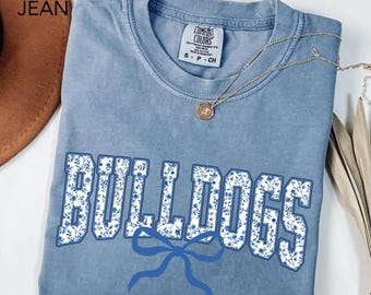 May include: A blue jean Comfort Colors t-shirt with the word "BULLDOGS" in white block letters filled with a blue floral pattern. A blue bow is below the word. A gold necklace with a pendant is on the shirt.