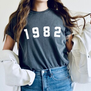 K&ouml;nnte beinhalten: Ein dunkelgraues T-Shirt mit dem Jahr 1982 in wei&szlig; auf der Vorderseite.