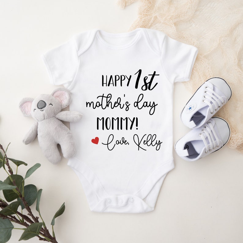May include: White baby onesie with the text "HAPPY 1st mother's day MOMMY! Love, Kelly" in black script. A red heart is next to the text "Love".
