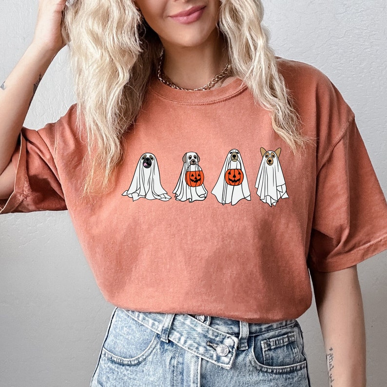 May include: A rust-colored t-shirt with a graphic of four dogs dressed as ghosts holding pumpkins. The dogs are a pug, a labrador, a corgi, and a chihuahua.