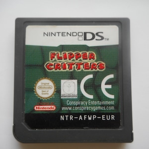 Könnte beinhalten: Eine Nintendo DS-Spielkassette für "Flipper Critters". Der Spieletitel ist in roter und gelber Schrift auf grünem Hintergrund. Die Kassette hat das Nintendo DS-Logo und das Conspiracy Entertainment-Logo.
