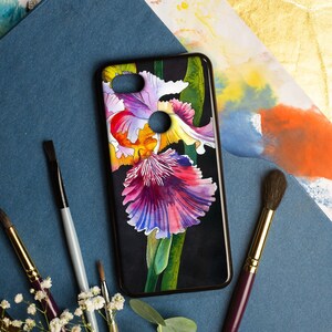 May include: Black phone case featuring a watercolor iris flower design in shades of purple, pink, yellow, and white. The case has a black border and is surrounded by paintbrushes and flowers.