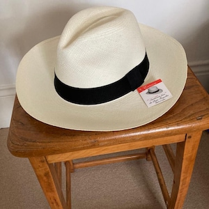 May include: A white Panama hat with a black ribbon and a red and white tag that reads "Elegance - Sombreros finos de Paja Toquilla".