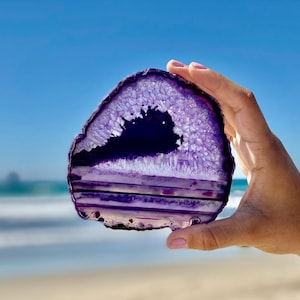 May include: A purple agate slice with white and dark purple banding. The agate is translucent and has a crystalline texture. The agate is being held in a hand against a blurred background of a beach and ocean.
